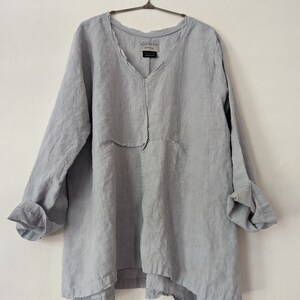 May include: A light gray linen tunic with a V-neck and long sleeves. The tunic has a relaxed fit and a layered hemline. The sleeves are rolled up at the cuffs. The brand name "breathe clothing" is visible on the neckline.