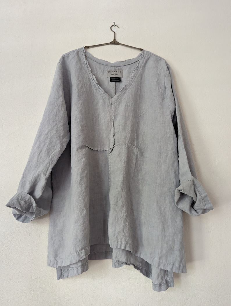 May include: A light gray linen tunic with a V-neck and long sleeves. The tunic has a relaxed fit and a layered hemline. The sleeves are rolled up at the cuffs. The brand name "breathe clothing" is visible on the neckline.