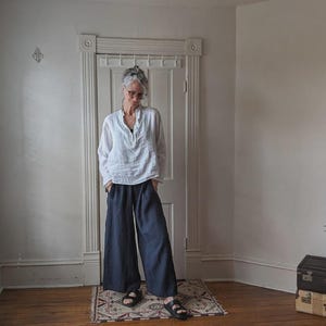 May include: A person wearing a white, long-sleeved shirt and navy blue wide-leg pants. The person is wearing black sandals. The room has a window with blinds and a door. A small rug is on the floor.