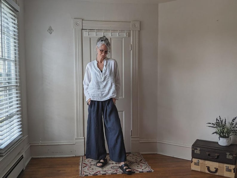 May include: A person wearing a white, long-sleeved shirt and navy blue wide-leg pants. The person is wearing black sandals. The room has a window with blinds and a door. A small rug is on the floor.