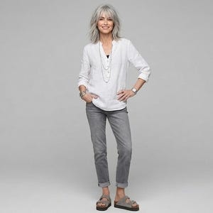 May include: A woman wearing a white long-sleeve shirt, gray jeans, and sandals. The shirt has a v-neck and is partially unbuttoned. The jeans are cuffed at the ankles. She is wearing a watch and jewelry.