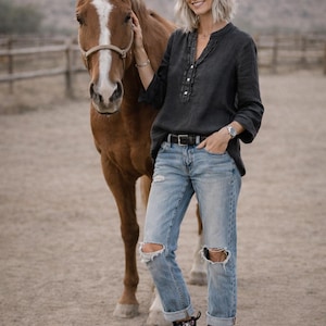 May include: A woman in a dark gray button-down shirt, light blue ripped jeans, and black floral boots stands next to a brown horse. The horse has a white blaze on its face. The background is a dusty outdoor setting.