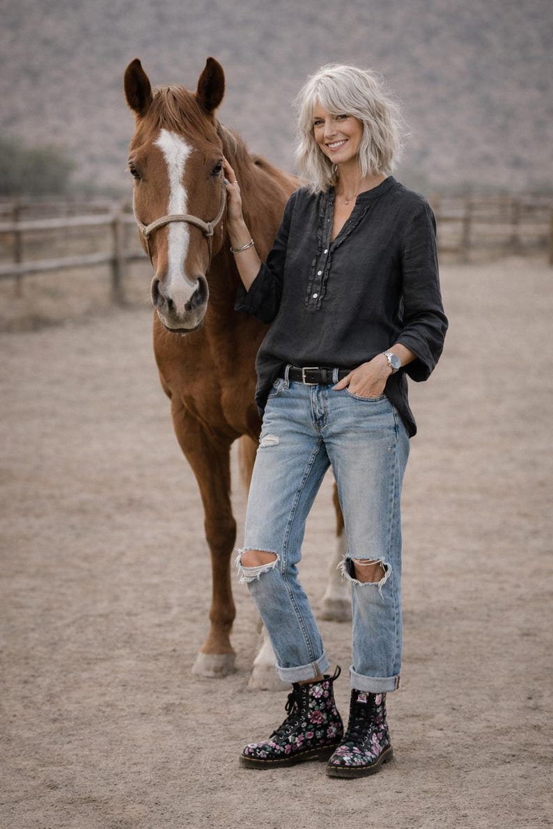 May include: A woman in a dark gray button-down shirt, light blue ripped jeans, and black floral boots stands next to a brown horse. The horse has a white blaze on its face. The background is a dusty outdoor setting.