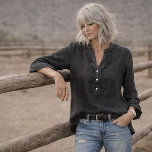 以下が含まれることがあります： フリルネックとボタンダウンフロントが特徴のダークグレーのリネンブラウス。ブラウスはブルージーンズと黒いベルトと組み合わせています。モデルは田舎の木製のフェンスに寄りかかっています。ブラウスは長袖です。