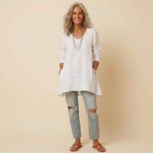 May include: A woman wearing a white tunic top with a V-neck and long sleeves, paired with light-wash, distressed jeans. The outfit is accessorized with a layered necklace and sandals. The background is a solid beige color.