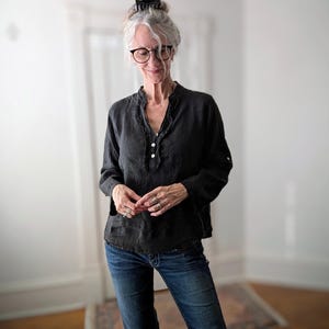 May include: A black long-sleeve blouse with a ruffled neckline and three white buttons. The blouse is paired with blue denim jeans. The woman in the image is wearing glasses and rings.