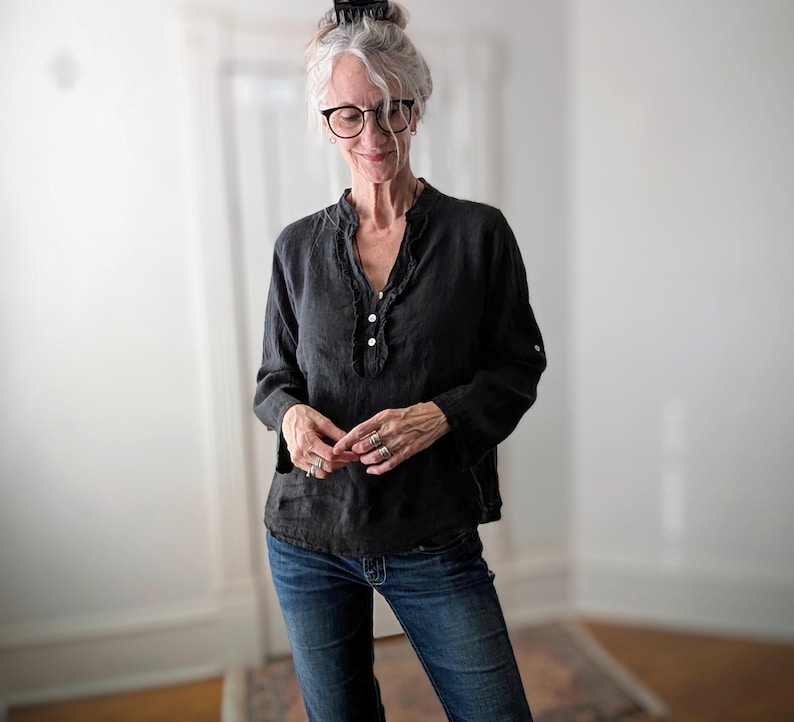 May include: A black long-sleeve blouse with a ruffled neckline and three white buttons. The blouse is paired with blue denim jeans. The woman in the image is wearing glasses and rings.