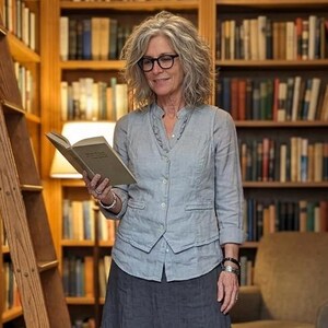 May include: A woman in a light blue vest and gray skirt stands in a library, holding an open book. Wooden bookshelves filled with books and a wooden ladder are in the background. A lamp provides soft lighting.