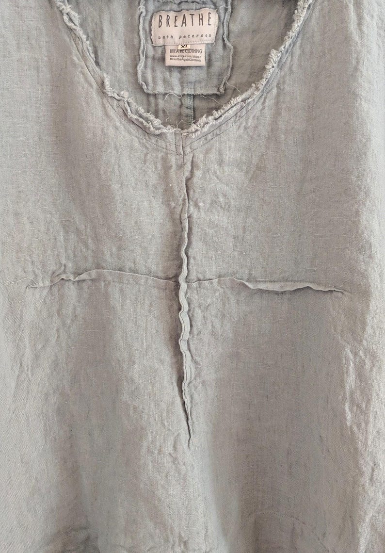 May include: Light blue linen top with a frayed neckline and seams. The top features a V-neck and a small tag that reads "BREATHE". The fabric has a slightly wrinkled texture.