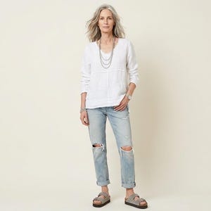 May include: A woman wearing a white long-sleeved top, light blue ripped jeans, and sandals. She is also wearing a silver necklace and a watch. The outfit is casual and comfortable.