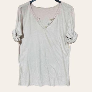 May include: A light beige henley shirt with a V-neck and three buttons. The long sleeves are rolled up. The shirt is hanging on a black hanger against a light background.