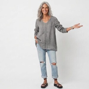 May include: A woman wearing a gray linen top with a V-neck and long sleeves, paired with light blue ripped jeans and black sandals. The outfit is casual and comfortable, suitable for everyday wear.