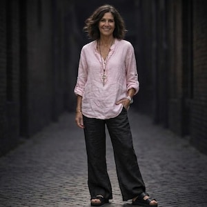 May include: A woman wearing a light pink linen shirt and dark gray pants stands in a cobblestone alley. She wears a necklace, bracelet, and sandals. The alley is flanked by dark brick walls.