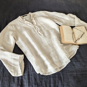 May include: A light-colored linen shirt with long sleeves and a ruffled neckline. The shirt is laid on a dark blue surface next to an open book with eyeglasses.