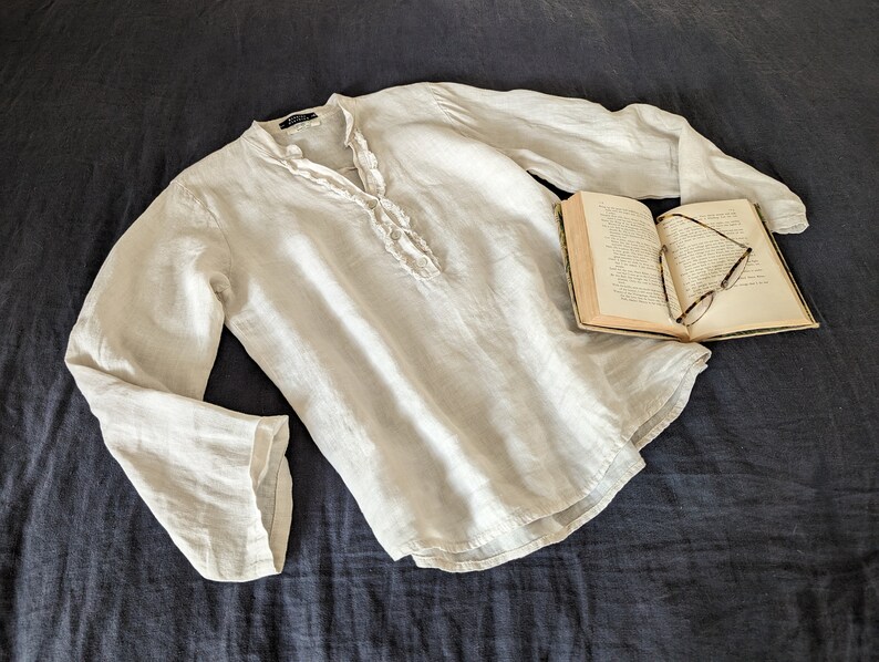 May include: A light-colored linen shirt with long sleeves and a ruffled neckline. The shirt is laid on a dark blue surface next to an open book with eyeglasses.