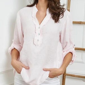 May include: A light pink linen blouse with a ruffled neckline and three small buttons. The long sleeves are rolled up to the elbows. The blouse has a relaxed fit and is made of a lightweight fabric. The model is wearing white pants.
