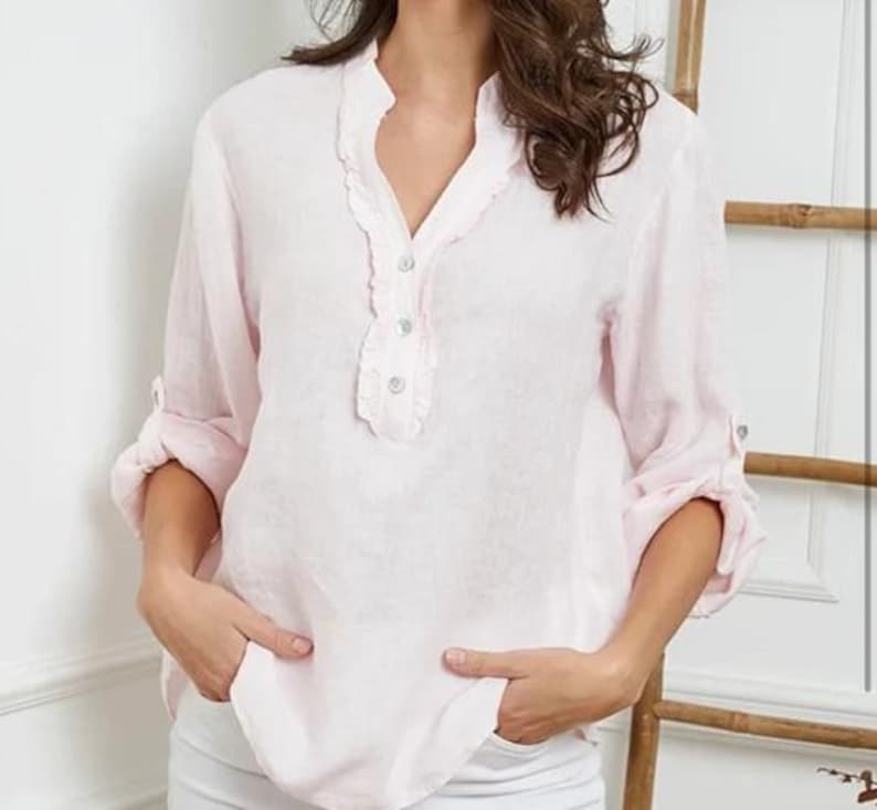 May include: A light pink linen blouse with a ruffled neckline and three small buttons. The long sleeves are rolled up to the elbows. The blouse has a relaxed fit and is made of a lightweight fabric. The model is wearing white pants.