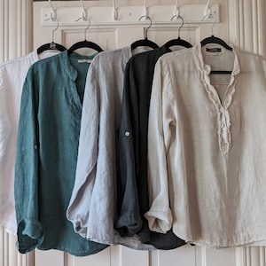 May include: Five linen shirts in white, teal, light gray, dark gray, and beige hang on black hangers. Each shirt has long sleeves and a mandarin collar. The beige shirt has a ruffled placket. The shirts are displayed against a white door.
