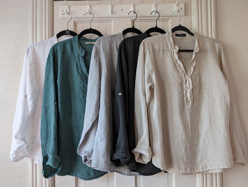 May include: Five linen shirts in white, teal, light gray, dark gray, and beige hang on black hangers. Each shirt has long sleeves and a mandarin collar. The beige shirt has a ruffled placket. The shirts are displayed against a white door.
