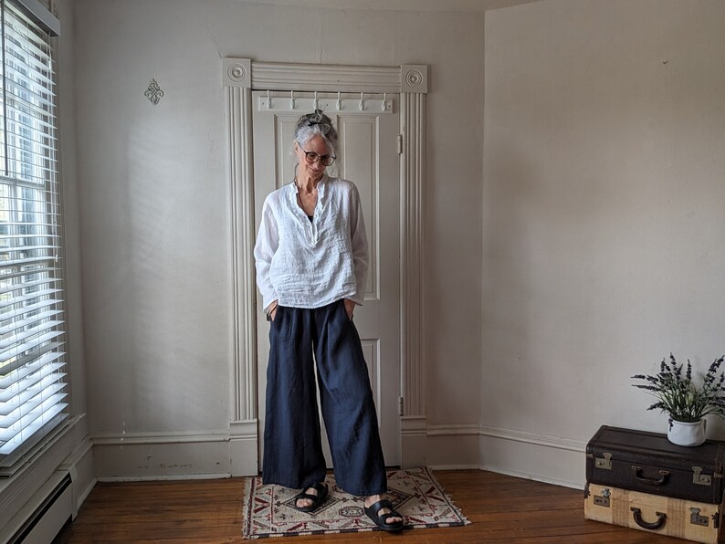 May include: A person wearing a white linen top and navy blue wide-leg pants. The outfit is paired with black sandals. The clothing is casual and comfortable. The person is standing in a room with a door and window.