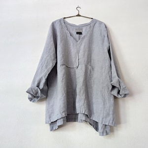 May include: A light gray linen tunic with a V-neck and long sleeves. The sleeves are rolled up at the elbows. The tunic hangs on a metal hanger against a white wall. The tunic has a relaxed, oversized fit.