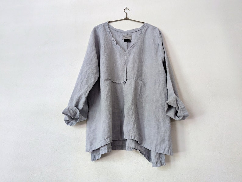 May include: A light gray linen tunic with a V-neck and long sleeves. The sleeves are rolled up at the elbows. The tunic hangs on a metal hanger against a white wall. The tunic has a relaxed, oversized fit.
