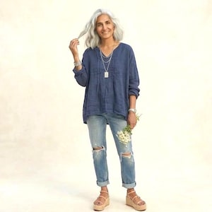 May include: A woman wearing a blue long-sleeve top, light-wash ripped jeans, and platform sandals. She holds a small bouquet of white flowers. The outfit is accessorized with silver jewelry. The background is a solid, light beige color.