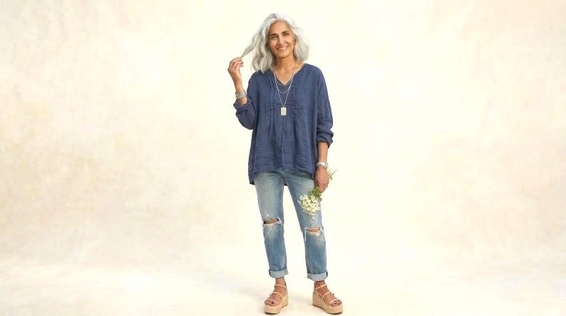 May include: A woman wearing a blue long-sleeve top, light-wash ripped jeans, and platform sandals. She holds a small bouquet of white flowers. The outfit is accessorized with silver jewelry. The background is a solid, light beige color.
