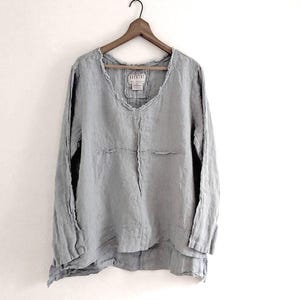 May include: A light gray long-sleeve linen shirt with a scoop neck, hanging on a wooden hanger. The shirt has a relaxed fit and a layered hemline. The fabric appears slightly textured.