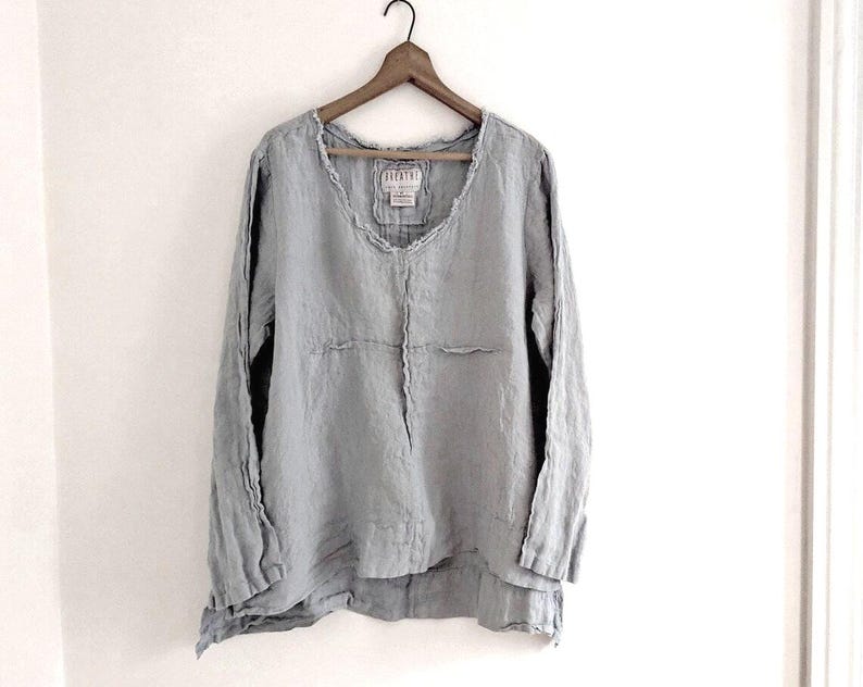 May include: A light gray long-sleeve linen shirt with a scoop neck, hanging on a wooden hanger. The shirt has a relaxed fit and a layered hemline. The fabric appears slightly textured.