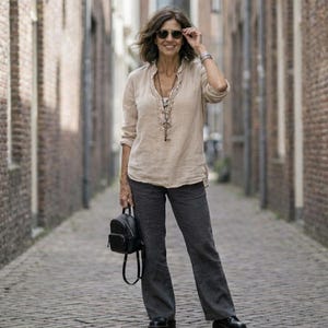 May include: A woman wearing sunglasses, a beige long-sleeve shirt, dark gray pants, and black shoes stands in a brick-lined alley. She carries a small black backpack and wears a necklace with a key pendant. The alley is paved with cobblestones.