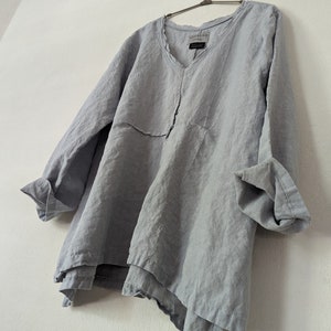 May include: A light blue linen tunic with a V-neck and long sleeves. The tunic has a layered, asymmetrical hemline and is hanging on a metal hanger. The fabric appears to be lightweight and slightly wrinkled.