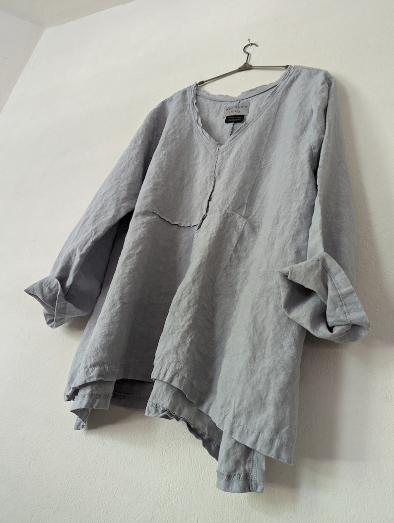May include: A light blue linen tunic with a V-neck and long sleeves. The tunic has a layered, asymmetrical hemline and is hanging on a metal hanger. The fabric appears to be lightweight and slightly wrinkled.