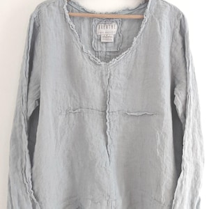 May include: A light blue linen long-sleeve shirt with a V-neck and frayed edges, hanging on a wooden hanger. The shirt has a relaxed fit and a layered hem. The label reads "BREATHE".