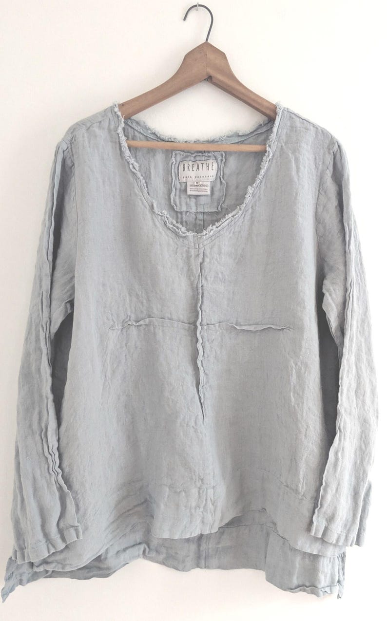 May include: A light blue linen long-sleeve shirt with a V-neck and frayed edges, hanging on a wooden hanger. The shirt has a relaxed fit and a layered hem. The label reads "BREATHE".