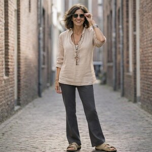 May include: A woman wearing a beige linen shirt, dark gray pants, and sandals stands in a brick-lined alley. She wears sunglasses and a necklace. The outfit is casual and suitable for warm weather.