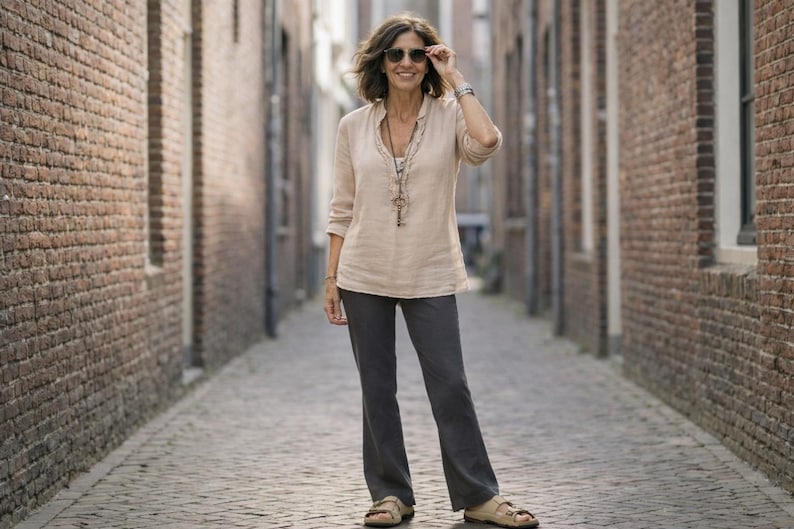 May include: A woman wearing a beige linen shirt, dark gray pants, and sandals stands in a brick-lined alley. She wears sunglasses and a necklace. The outfit is casual and suitable for warm weather.