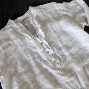 May include: A white linen shirt with long sleeves. The shirt features a ruffled collar and a button-down placket with three buttons. The fabric has a slightly textured appearance. The shirt is laid flat on a dark surface.
