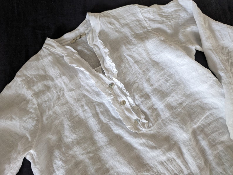 May include: A white linen shirt with long sleeves. The shirt features a ruffled collar and a button-down placket with three buttons. The fabric has a slightly textured appearance. The shirt is laid flat on a dark surface.