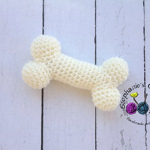 May include: A cream-coloured, crocheted dog bone toy. The bone is made of soft yarn and has a textured appearance. The toy is designed with rounded ends and a central shaft, resembling a real bone. The background is a distressed white wooden surface.