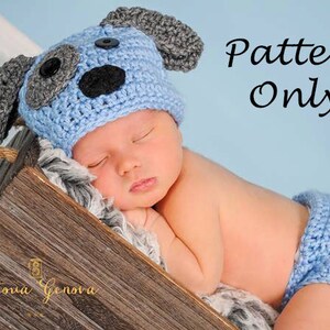 Puede incluir: Un conjunto de gorro y pañal de crochet azul con orejas de perro grises y una nariz negra. El conjunto está sobre una superficie de madera con una manta blanca y esponjosa.