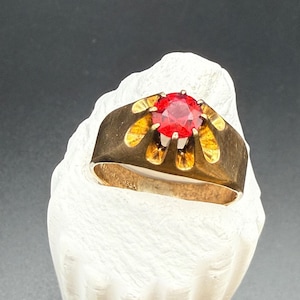May include: A gold ring featuring a round, faceted, vibrant red gemstone set in a wide, polished band. The setting has a distinct petal-like design surrounding the stone. The interior of the band is engraved with the text 14K GOLDSHELL.