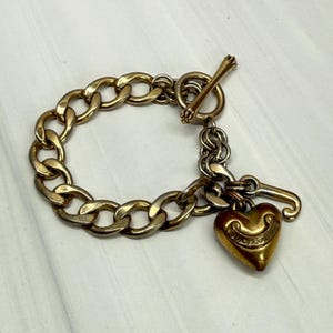May include: A gold-tone metal chain link bracelet featuring a toggle clasp and a heart-shaped charm. The charm is embossed with the text Juicy Couture and includes a small J-shaped accent piece attached to the same jump ring.