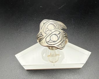 May include: A silver ring featuring an engraved abstract design with curved lines and circular motifs. The metal surface has a polished finish with darkened recessed areas that highlight the intricate patterns across the top of the band.