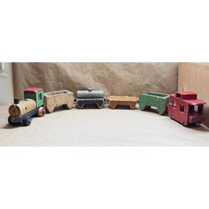 May include: A wooden toy train set consisting of a locomotive with a green cab, a natural wood cargo car, a gray tank car, a flatbed car, a green open-top car, and a red caboose. Each car features small wheels and a weathered, painted finish.
