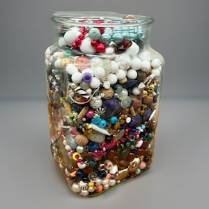 May include: A clear glass jar filled with a variety of beads, shells, and small decorative items. The contents include white, red, blue, and brown beads, along with other colorful elements. The jar is square with rounded edges.