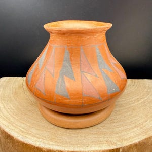 Könnte beinhalten: Eine terrakottafarbene Keramikvase mit einem geometrischen Design in Grau und Lachs. Die Vase hat einen abgerundeten Körper und einen leicht ausgestellten Rand und steht auf einem passenden kreisförmigen Sockel. Das Design zeigt sich wiederholende Dreiecksformen.