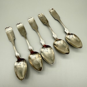 May include: Five antique silver spoons arranged diagonally. Each spoon features a heart-shaped design at the end of the handle and a rounded bowl. The spoons have a tarnished appearance, suggesting age and use.