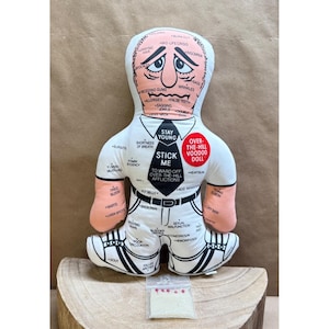 May include: A novelty voodoo doll with a sad face, labeled with various ailments. The doll is white with black accents, wearing a tie that says "Stick Me". A small bag of pins sits below the doll.