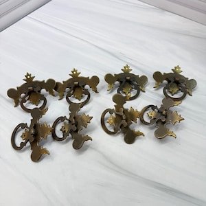 May include: A set of six brass-toned metal drawer pulls featuring ornate, symmetrical backplates with intricate cut-out details and circular drop handles. Each piece displays a weathered, aged finish with visible mounting points.